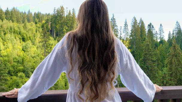 Mooie jonge vrouw in badjas staat op het terras of balkon van het hotel en kijkt uit op het dennenbos en de bergen. Begrip toerisme en vakantie in de bergen in de zomer - Foto, afbeelding