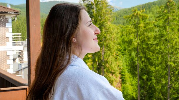 Portrét krásné usměvavé ženy v županu těší pohled na hory a les ze svého hotelového pokoje. Koncept cestovního ruchu a dovolené v horách v létě - Fotografie, Obrázek