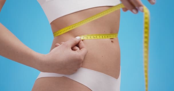 Jeune dame élancée en sous-vêtements mesurant sa taille avec mètre, vue latérale, fond studio bleu - Séquence, vidéo