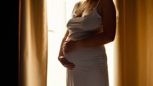 Close-up van mooie zwangere vrouw in nachtjapon staan tegen felle zon schijnt door groot raam en streelde haar grote buik. Concept van gelukkige zwangerschap en het verwachten van een baby - Foto, afbeelding