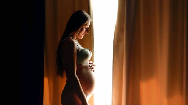 Beautiful pregnant woman in lingeri opens curtain at big window and looking on her big belly.Concept of happy pregnancy and baby anticipation - Photo, Image