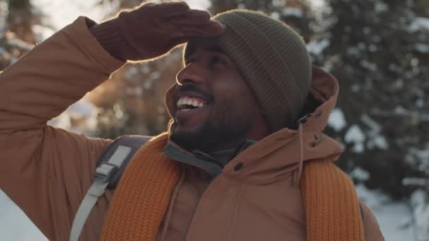 Αργή κίνηση γκρο πλαν του χαρούμενου μαύρου άνδρα κοιτάζοντας γύρω απολαμβάνοντας χιόνι το χειμώνα στέκεται στο γραφικό δάσος χαμογελώντας γενικά - Πλάνα, βίντεο