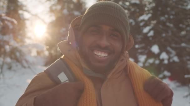 Zbliżenie portret uśmiechniętego czarnego mężczyzny z plecakiem stojącego w zimowym lesie z mnóstwem śniegu patrzącego szczęśliwie w kamerę - Materiał filmowy, wideo