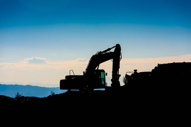Backlit silhouette of an excavator on top of a hill with unfocused sky background. - Photo, Image