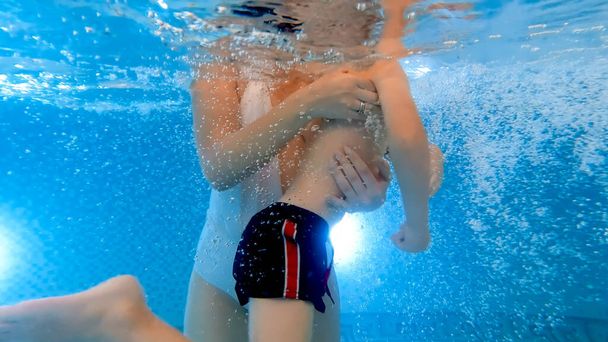 水中では、水泳プールで泳ぐ方法を学ぶ彼女の小さな息子を支える若い母親を撮影しました。家族の健康とスポーツの概念. - 写真・画像