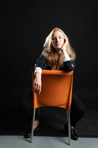 A young girl is sitting on an orange chair. Smiling with charismatic appearance, concept of webinar activity, on a black background in the studio, in pedjak business woman, blonde - Fotoğraf, Görsel