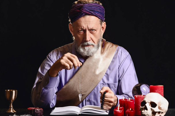 Male fortune teller with pendulum and book at table on dark background - Photo, Image