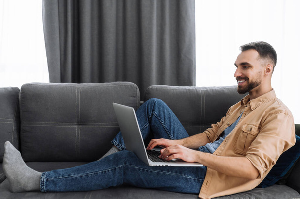 Caucasian male freelancer sits on the sofa, chilling at home. Bearded guy in casual wear use a laptop, watching films or funny videos, or working from home and smiling - 写真・画像