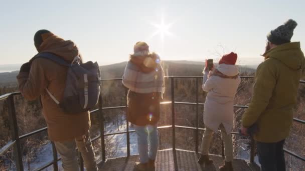 Middellange achteruitkijkbeelden van een groep toeristen die op het uitkijkpunt staan en foto 's maken van prachtige taferelen met besneeuwd bos en bergen in het zonlicht - Video