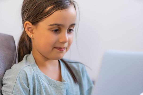 Ένα κοριτσάκι σε σχολική ηλικία σπουδάζει στο σπίτι. Ένα κορίτσι παρακολουθεί ένα μάθημα βίντεο σε μια οθόνη laptop και γράφει εργασίες σε ένα σημειωματάριο. - Φωτογραφία, εικόνα