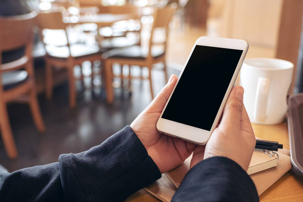 Mockup kuva käsissä tilalla ja käyttämällä valkoista matkapuhelinta tyhjällä näytöllä kannettavan ja kahvikupin puinen pöytä kahvilassa - Valokuva, kuva