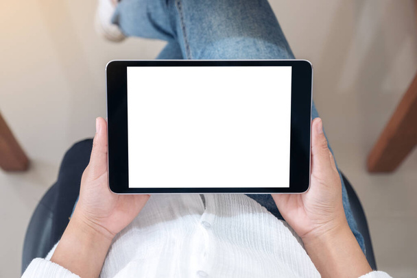 Imagen de maqueta de vista superior de una mujer sosteniendo una tableta negra con pantalla blanca en blanco horizontalmente mientras está sentada en una silla - Foto, Imagen