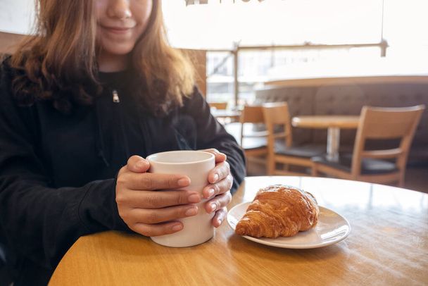 Eine Frau hält eine Kaffeetasse mit einem Stück Croissant in einem weißen Teller auf einem Holztisch im Café - Foto, Bild