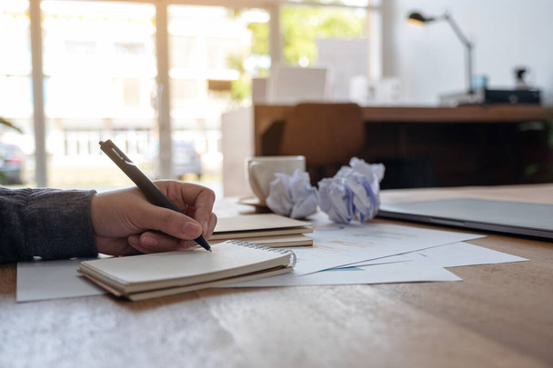 Imagen de cerca de una mujer de negocios que trabaja y escribe en un cuaderno blanco en blanco con papeles arruinados y computadora portátil en la mesa en la oficina - Foto, Imagen