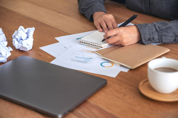 Close-up beeld van een zakenvrouw werken en opschrijven op een wit blanco notitieboekje met verknipte papieren en laptop op tafel in het kantoor - Foto, afbeelding