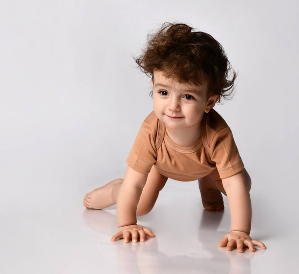 Charming dark-haired curly baby boy crawls on all fours on a gray background. - Φωτογραφία, εικόνα