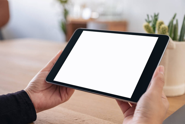 Mockup afbeelding van handen houden zwarte tablet pc met blanco wit scherm horizontaal tijdens het zitten in het kantoor - Foto, afbeelding