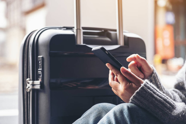 Closeup image of a woman sitting and using mobile phone with a black baggage for traveling - Photo, Image