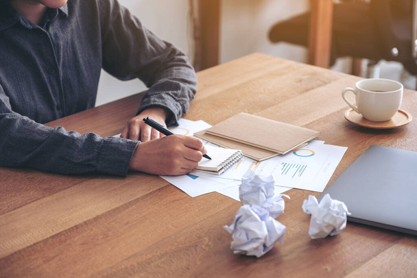 Close-up beeld van een zakenvrouw werken en opschrijven op een wit blanco notitieboekje met verknipte papieren en laptop op tafel in het kantoor - Foto, afbeelding