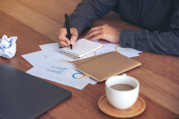 Detailní obrázek podnikatelky pracující a zapisující si na bílý prázdný zápisník se zašmodrchanými papíry a notebookem na stole v kanceláři - Fotografie, Obrázek