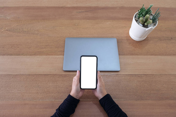 Вид зверху макет зображення рук, що тримають порожній білий екран мобільний телефон і ноутбук на дерев'яному столі в офісі
 - Фото, зображення