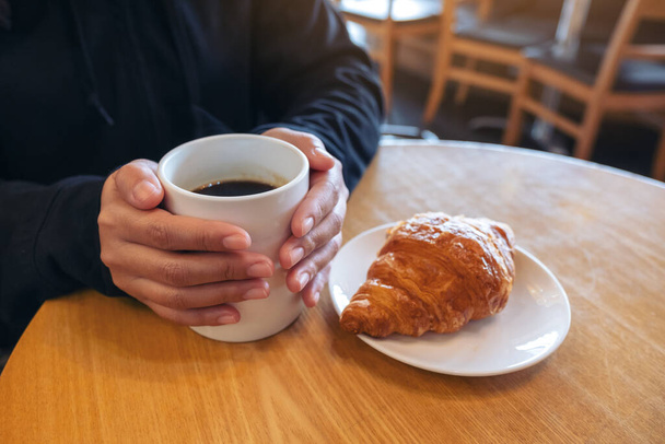 Kobieta trzymająca filiżankę kawy z kawałkiem rogalika w białym talerzu na drewnianym stole w kawiarni - Zdjęcie, obraz