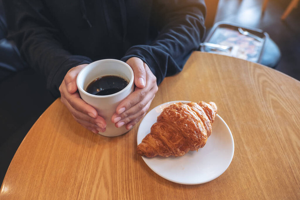 Eine Frau hält eine Kaffeetasse mit einem Stück Croissant in einem weißen Teller auf einem Holztisch im Café - Foto, Bild
