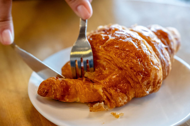 Szoros kép a kezek vágás egy darab croissant késsel és villával reggelire a fa asztalon  - Fotó, kép