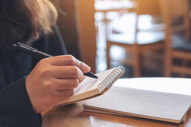 Imagen de primer plano de una mano sosteniendo y escribiendo en cuaderno en blanco en la mesa en la cafetería - Foto, Imagen