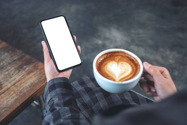 Mockup image of hands holding black mobile phone with blank screen while drinking coffee in cafe - Photo, Image