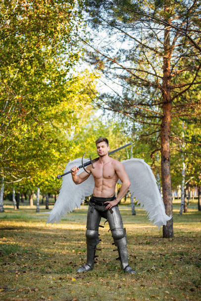 A young angel, a bodybuilder in plate armor on his legs with wings behind his back, holds in his hands a two-handed sword. - Фото, зображення