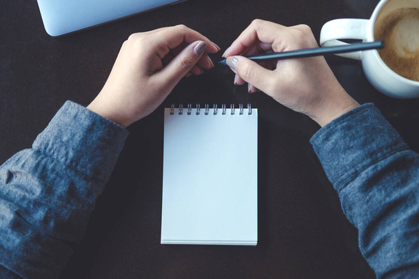 Widok z góry obraz dłoni kobiety trzymającej ołówek do pisania na pustym notebooku z laptopem i filiżanką kawy na stole  - Zdjęcie, obraz