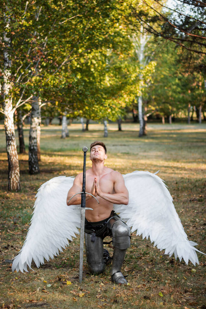 Portrait of a warrior angel with wings behind him, with a naked torso and in plate armor on his feet, sitting on one knee next to a two-handed sword and clasped hands in prayer. - Фото, зображення