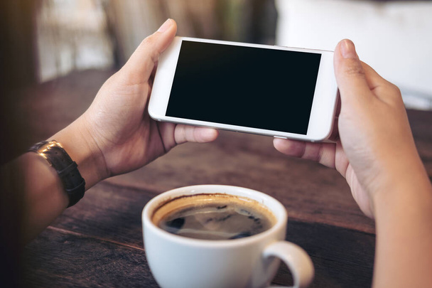 Imagen burlona de manos sosteniendo teléfono móvil blanco con pantalla negra en blanco para ver y jugar juegos con una taza de café en la mesa de madera en la cafetería - Foto, Imagen