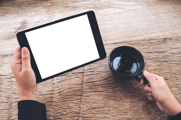 Mockup image of hands holding black tablet pc with blank white screen and coffee cup on vintage wooden table in cafe  - Zdjęcie, obraz