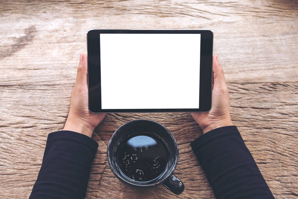 Imagem de maquete de vista superior de mãos segurando tablet preto pc com tela branca em branco e xícara de café na mesa de madeira vintage no café  - Foto, Imagem
