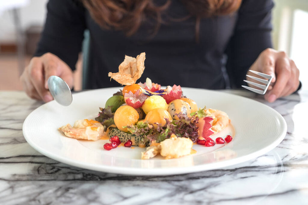 A woman holding spoon and fork and preparing to eat mixed fruits salad with fried shrimps on the table in restaurant - Photo, Image