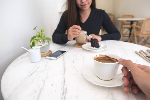 Imagem de close-up de uma mão segurando xícara de café e uma mulher comendo lanche na mesa de vidro no café moderno - Foto, Imagem