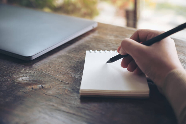 Nahaufnahme des Schreibens von Hand auf ein leeres Notizbuch mit Laptop auf Holztisch  - Foto, Bild