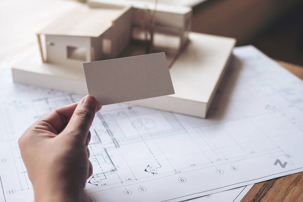 Une main tenant et montrant une carte de visite vide avec un modèle d'architecture et papier dessin de magasin sur la table dans le bureau  - Photo, image