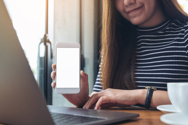 Mockup-Bild einer Frau, die ein weißes Handy mit leerem Bildschirm auf dem Tisch hält und zeigt, während sie Laptop benutzt - Foto, Bild