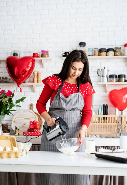 Valentijnsdag. Vrouw in rode jurk en grijze schort die Valentijnskoekjes maakt in de keuken - Foto, afbeelding