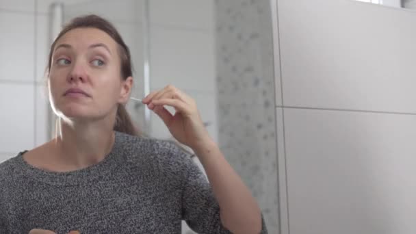 Kobieta czyści uszy pączkami bawełny rano przed lustrem - Materiał filmowy, wideo
