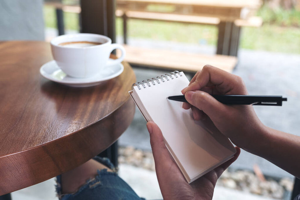 Image rapprochée des mains d'une femme écrivant sur un carnet blanc vierge avec tasse à café sur la table - Photo, image