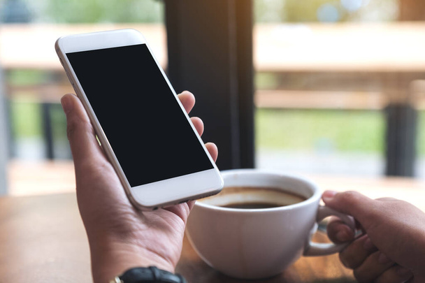 Mockup beeld van handen met witte mobiele telefoon met blanco zwart scherm tijdens het drinken van koffie in cafe - Foto, afbeelding
