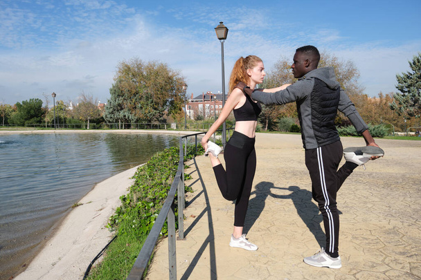 African black man and redhead caucasian woman stretching their legs in a park. Fitness and sport outdoor. - Photo, Image