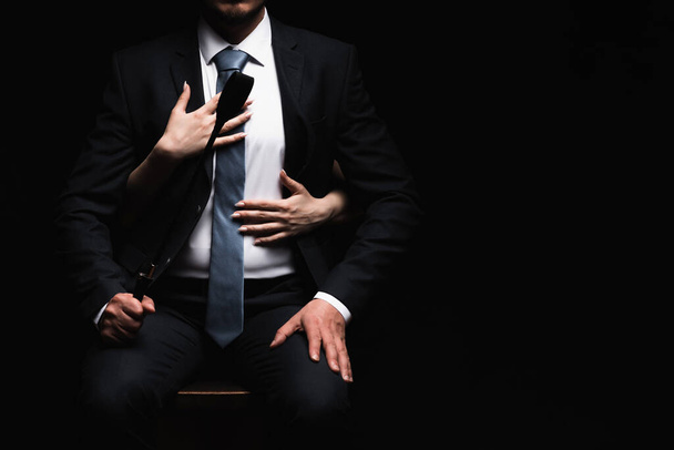 женские руки обнимают доминирующего мужчину в костюме с кожаным кнутом. Концепция БДСМ секса - Фото, изображение