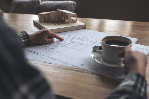 Een architect die werkt aan een architectuurmodel met tekenpapier tijdens het drinken van koffie op kantoor - Foto, afbeelding