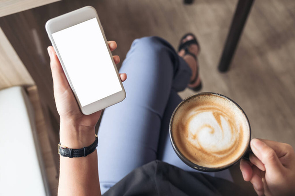 Top view makieta obrazu dłoni kobiety trzymającej biały telefon komórkowy z pustym ekranem pulpitu podczas picia kawy w kawiarni - Zdjęcie, obraz
