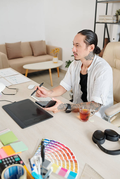 Ervaren ontwerper met smartphone in handen werkend op grafische tablet in zijn thuiskantoor - Foto, afbeelding